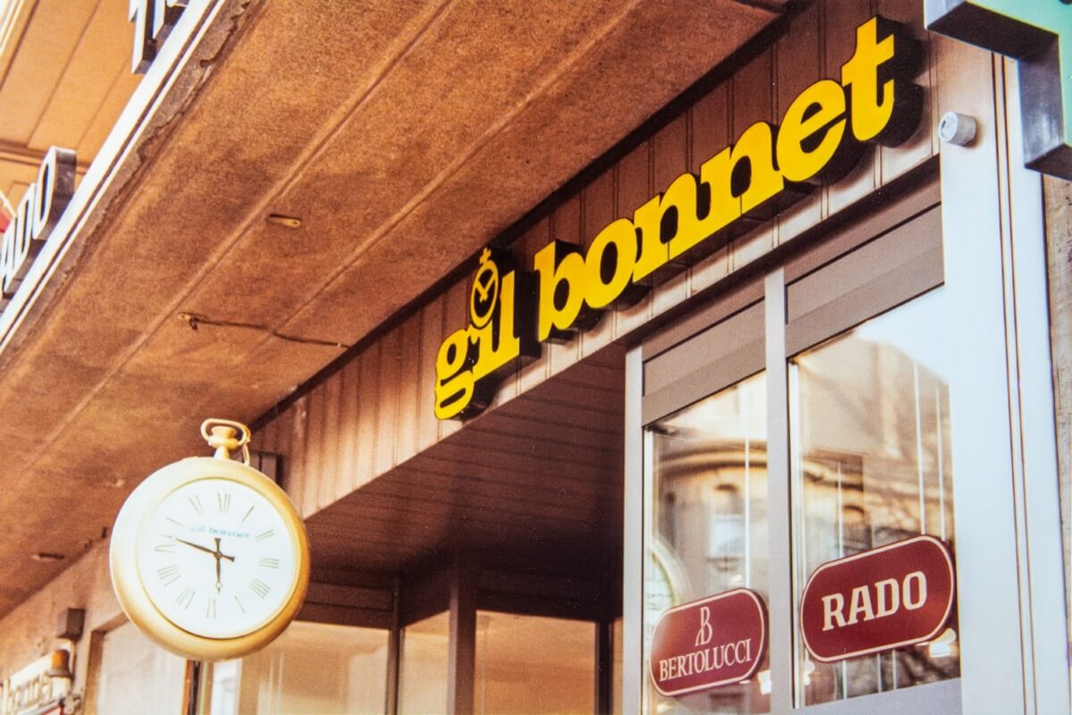 Gros plan sur l'enseigne jaune de Gil Bonnet et l’horloge suspendue en forme de montre à gousset.